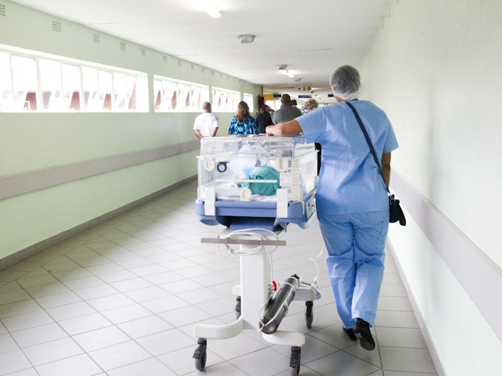 Clinician in blue scrubs walking a hospital corridor past neonatal equipment